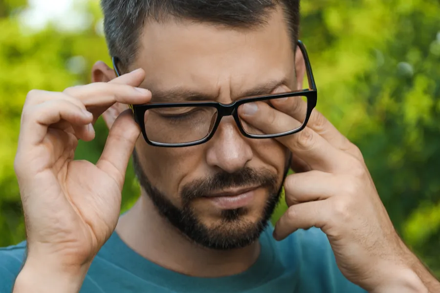 Allergic-Conjunctivitis-Clinic A man with a beard and glasses standing outside and wearing a blue t-shirt lifts his glasses and rubs his eyes due to allergy irritation. Get treatment for allergic conjunctivitis from Guerda Johnson, FNP-C in Tyler East Texas Area.
