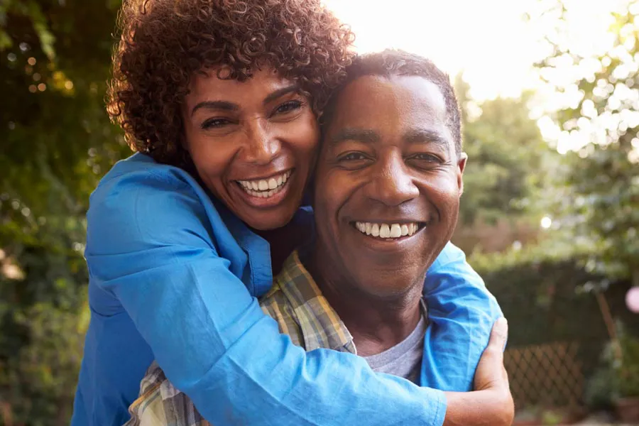 Autoimmune-Doctor A middle-aged African American couple outside, the woman embracing the man from behind pressing her cheek to his, both are smiling due to successful Autoimmune Disorder treatment from Guerda Johnson, FNP-C in Tyler East Texas Area.