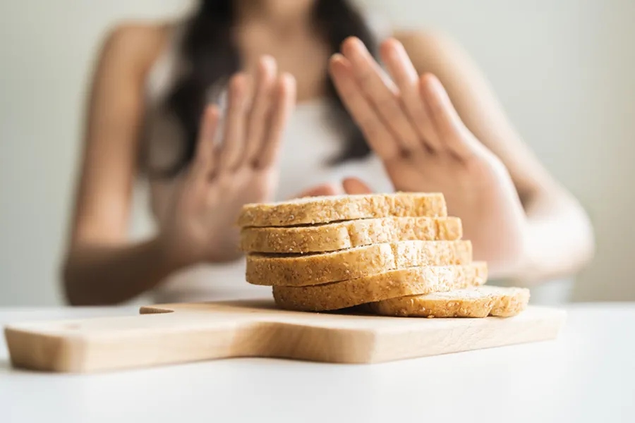 Celiac-Disease-Clinic Close up of a stack of bread slices on a cutting board with a woman's hands behind them, refusing to eat. Get treatment for Celiac Disease from Guerda Johnson, FNP-C in Tyler East Texas Area.