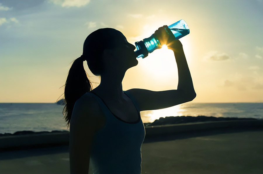 Chelation-and-Detoxification-Clinic A woman in exercise clothes drinking from a water bottle on the beach, with the water and sunset in the background. Get chelation and detoxification treatment from Guerda Johnson, FNP-C in Tyler East Texas Area.