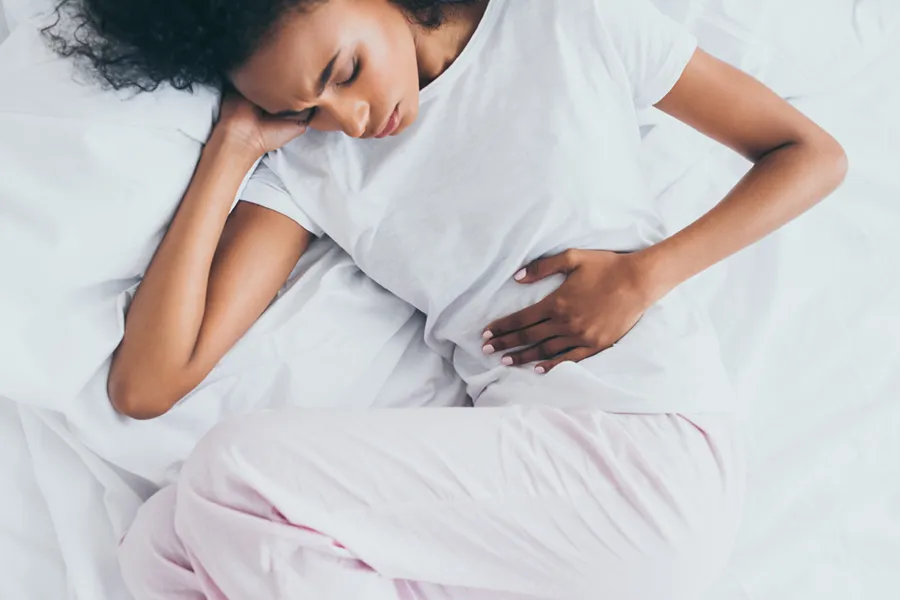 Crohns-Disease-Clinic A dark-skinned woman in white clothes lays on her bed clutching her middle before getting treatment for Crohn's Disease from Guerda Johnson, FNP-C in Tyler East Texas Area.