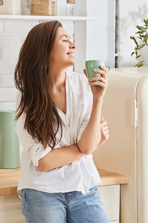 Crohns-Disease-Treatment A woman with a white blouse an jeans drinking tea in her kitchen, smiling after getting treatment for Crohn's Disease from Guerda Johnson, FNP-C in Tyler East Texas Area.