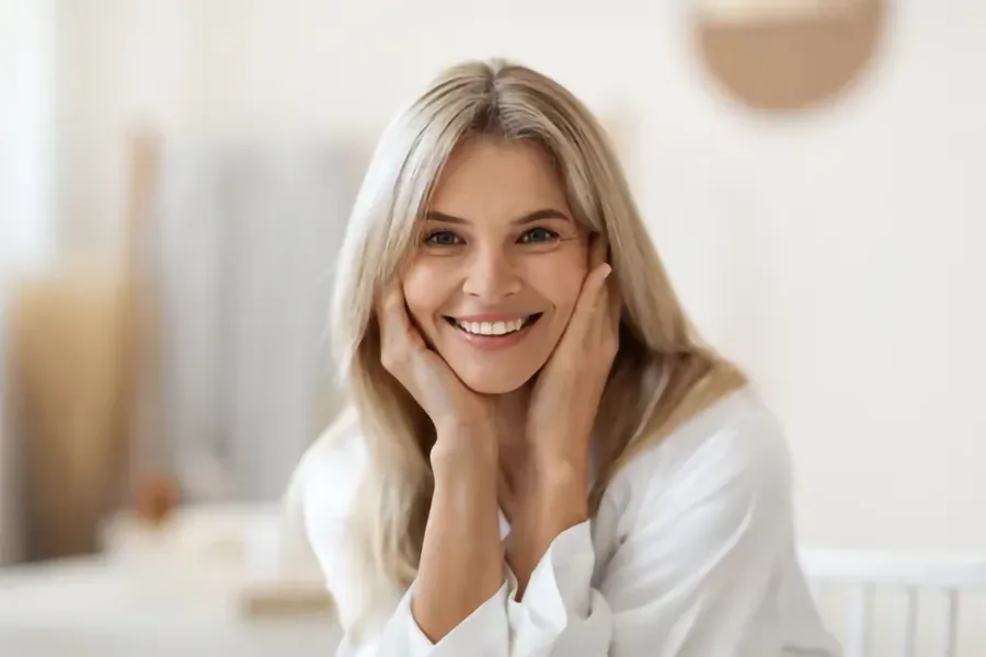 EmpowerRF-Vaginal-Rejuvenation-Clinic A cheerful middle-aged Caucasian woman with long, flowing blonde hair, smiling warmly while seated in a bright, cozy room after receiving EmpowerRF Vaginal Rejuvenation treatment by Guerda Johnson, FNP-C in Tyler East Texas Area.