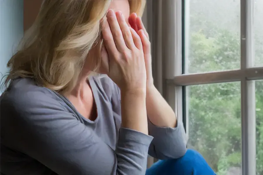 Graves-Disease-Clinic A blonde woman in a gray shirt sits in front of a window on a rainy day covering her eyes with her hands. Get treatment for Grave's Disease from Guerda Johnson, FNP-C in Tyler East Texas Area.
