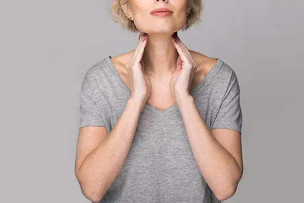 Hashimotos-Thyroiditis-Doctor An older blonde woman in a gray t-shirt holds her hands to her neck checking for signs of thyroid problems. Get help for Hashimoto's Thyroiditis from Guerda Johnson, FNP-C in Tyler East Texas Area.