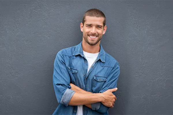 Hormones-for-Men-Doctor A man in a denim shirt stands smiling against a gray-blue wall, pleased with his testosterone hormone therapy from Guerda Johnson, FNP-C in Tyler East Texas Area.