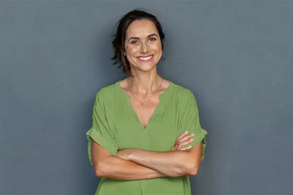 Hormones-for-Women-Doctor A woman in a green shirt stands smiling against a gray-blue wall, pleased with her hormone therapy from Guerda Johnson, FNP-C in Tyler East Texas Area.