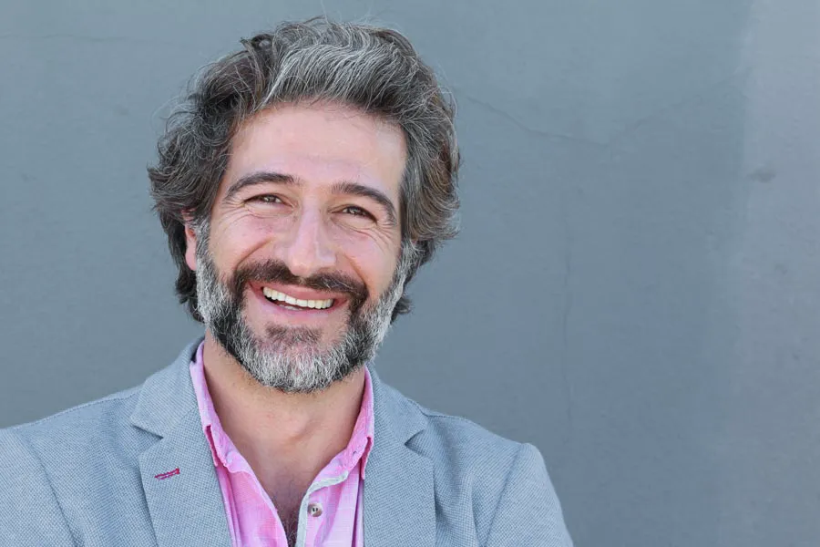 Hypogonadism-Clinic A smiling man with salt and pepper hair and beard wearing a pink shirt and gray blazer stands agains a gray wall. Get hypogonadism treatment from Guerda Johnson, FNP-C in Tyler East Texas Area.