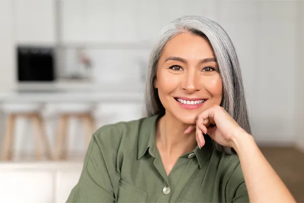 Menopause-Clinic An asian woman with long gray hair and a green shirt sitting and smiling on her couch, happy with her menopause care from [NAME} in Tyler East Texas Area.