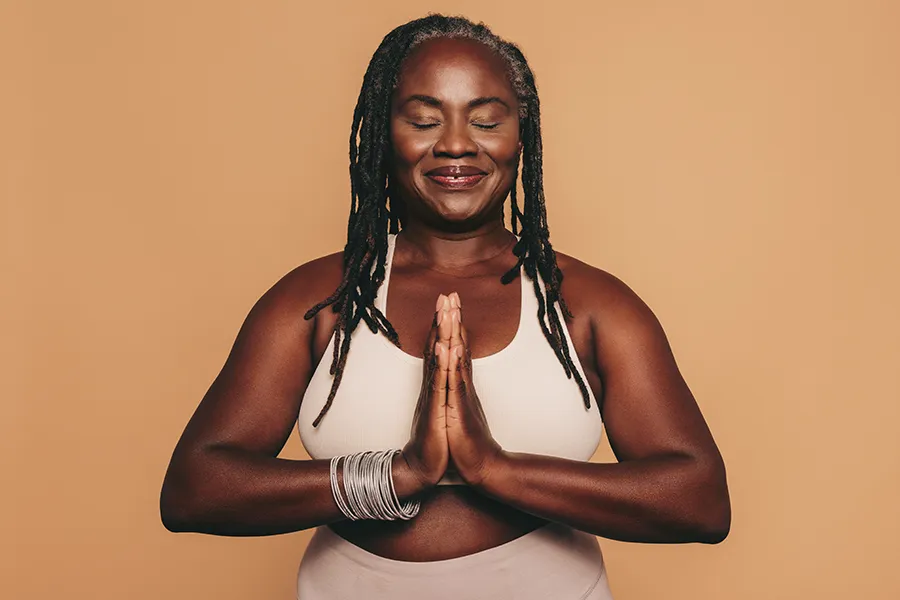 Obesity-Clinic An older African American woman confidently posing against a warm background smiling with hands in a prayer pose. Guerda Johnson, FNP-C treats obesity using functional medicine in Tyler East Texas Area.