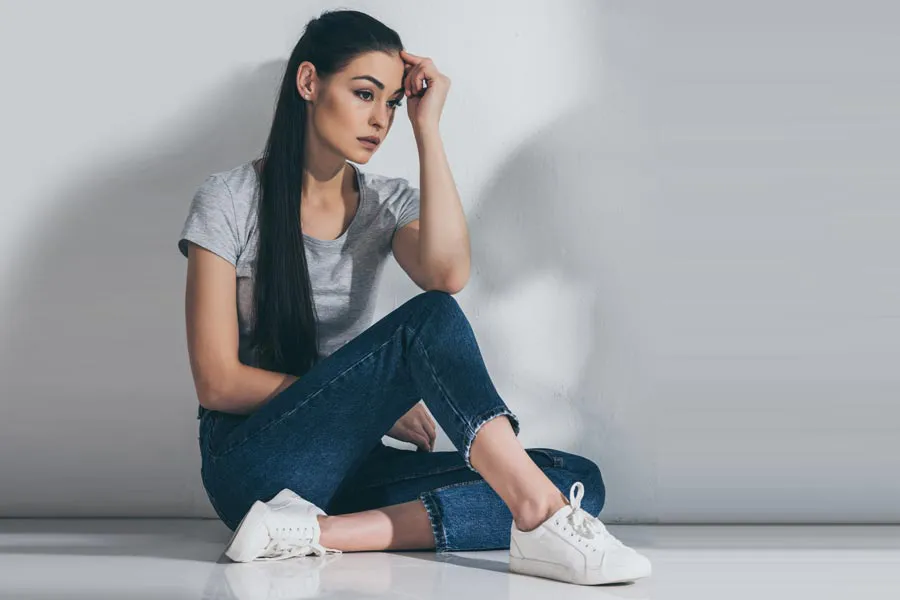 PCOS-Doctor A young brunette woman in a gray t-shirt and jeans sits against a gray wall with her head resting on her hand and a distressed expression. Get help for Polycystic Ovarian Syndrome (PCOS) from Guerda Johnson, FNP-C in Tyler East Texas Area.