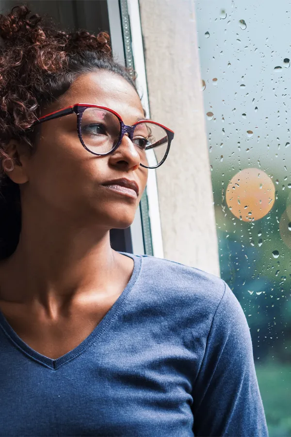 PMDD-Treatment A black woman with with a somber expression and glasses and a gray shirt stares out the window on a rainy day. Get treatment for Premenstrual Dysphoric Disorder from Guerda Johnson, FNP-C in Tyler East Texas Area.