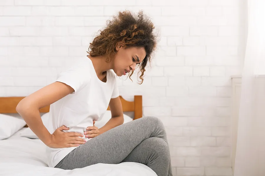 PMS-Clinic-2 A black woman sits on her bed holding her stomach in pain. Get treatment for PMS from Guerda Johnson, FNP-C in Tyler East Texas Area.
