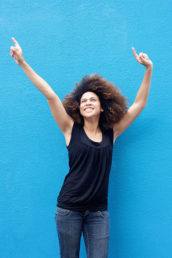 PMS-Treatment A woman in a dark blue tank top standing in front of a bright blue wall, raising her arms in celebration of relief from PMS from Guerda Johnson, FNP-C in Tyler East Texas Area.