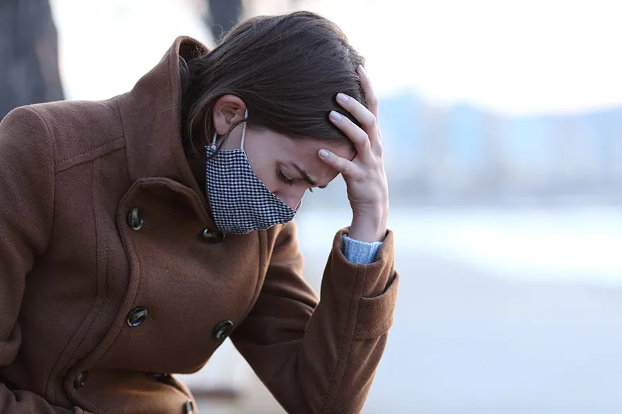 Post-Covid-Clinic A woman in a mask sits outside with her hand on her forehead in pain. Get long covid treatment from Guerda Johnson, FNP-C in Tyler East Texas Area.