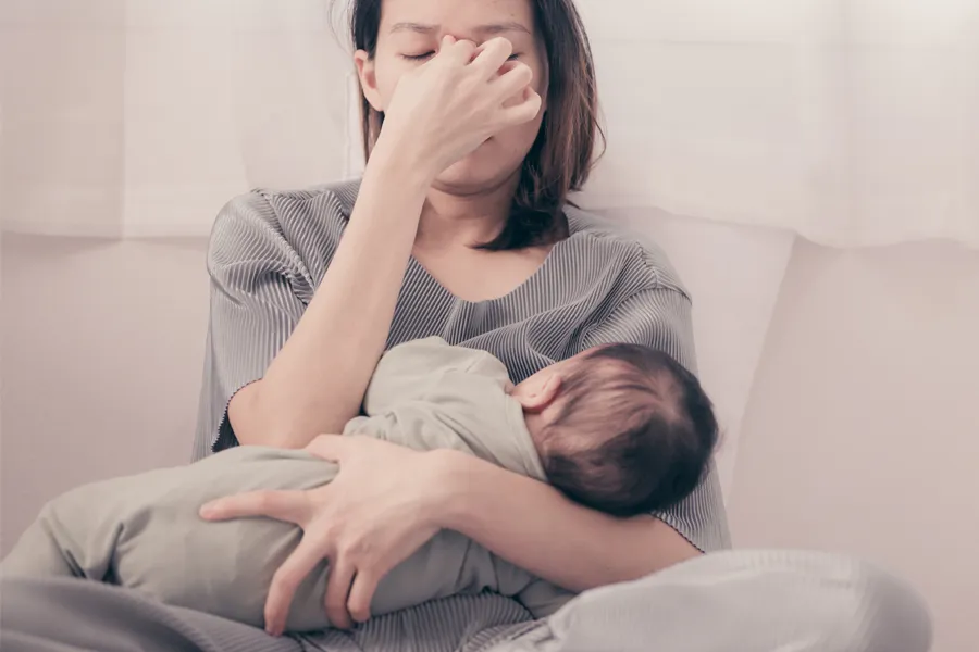 Postpartum-Hormone-Imbalance-Clinic A woman nursing an infant on couch rubs her eyes with exhaustion. Get treatment for postpartum hormone imbalance from Guerda Johnson, FNP-C in Tyler East Texas Area.