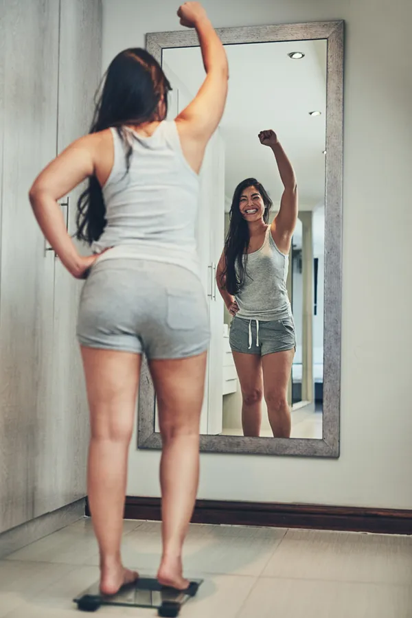 Tirzepatide-Treatment A woman in workout clothes stands on a scale looking in the mirror and pumping her hands in celebration of her successful Tirzepatide treatment from Guerda Johnson, FNP-C in Tyler East Texas Area.