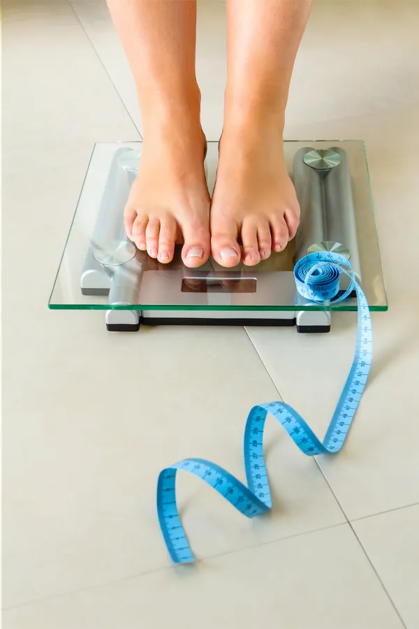 Weight-Loss-Resistance-Treatment Close-up of a woman's feet standing on a scale, with measuring tape by her toes, getting treatment for weight loss resistance from Guerda Johnson, FNP-C in Tyler East Texas Area.